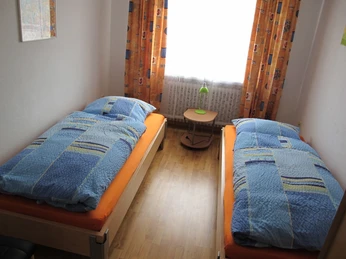 Schlafraum Zwei Einzelbetten mit blauen Bettdecken stehen in einem hellen Raum mit Holzfußboden und buntem Vorhang.