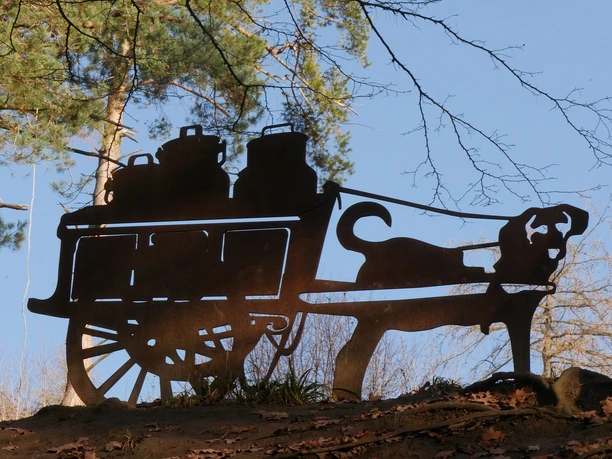 Skulptur eines Milchkarrens Metallskulptur eines Milchkarrens, gezogen von einem Hund. Eingerahmt von Bäumen und blauem Himmel.