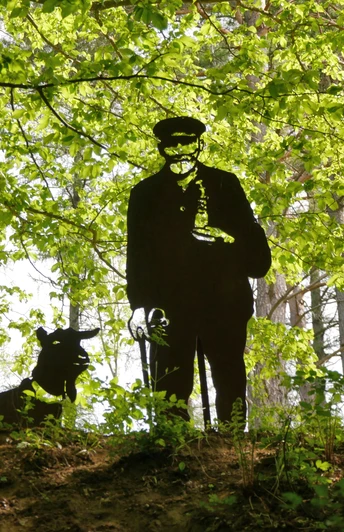 Eine dunkle Schäferskulptur steht neben einem Schäferhund im lichten Wald unter grünem Blätterdach.