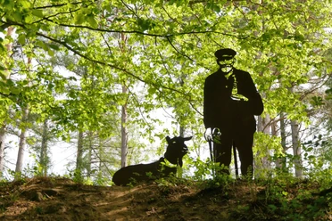 Schäferskulptur im Holweg Eine dunkle Schäferskulptur steht neben einem Schäferhund im lichten Wald unter grünem Blätterdach.