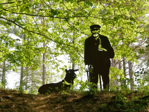 Schäferskulptur im Holweg Eine dunkle Schäferskulptur steht neben einem Schäferhund im lichten Wald unter grünem Blätterdach.