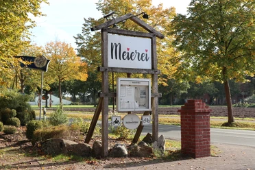 Meierei Verl Holzschild mit Aufschrift "Meierei" vor herbstlicher Allee, umgeben von Bäumen und Landschaft.