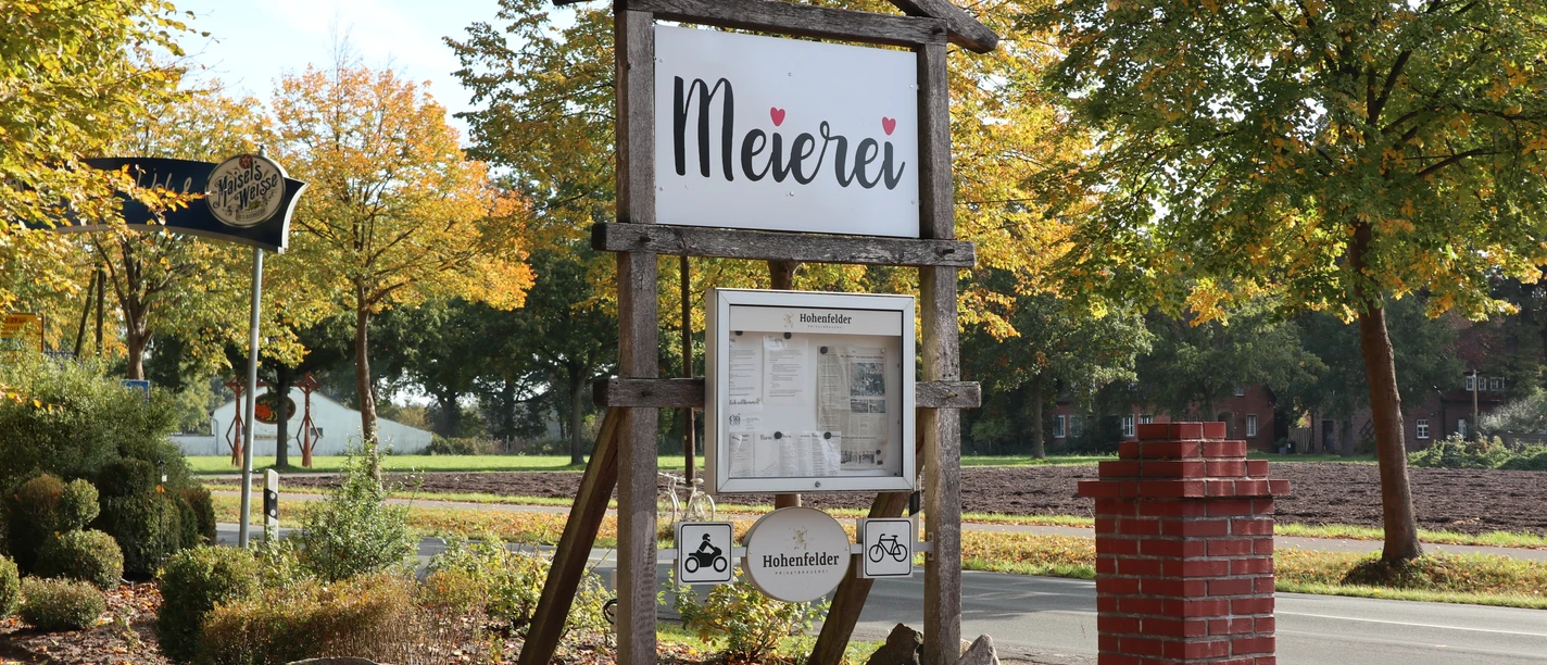 Meierei Verl Holzschild mit Aufschrift "Meierei" vor herbstlicher Allee, umgeben von Bäumen und Landschaft.