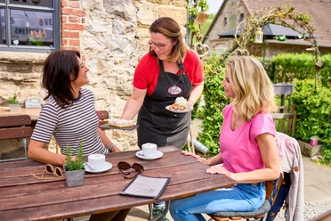 Café Herberge in Heiligenhaus Zwei Personen sitzen an einem Holztisch im Freien, während eine Kellnerin Kuchen serviert.