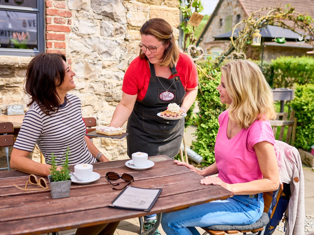 Café Herberge in Heiligenhaus Zwei Personen sitzen an einem Holztisch im Freien, während eine Kellnerin Kuchen serviert.