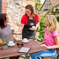 Café Herberge in Heiligenhaus Zwei Personen sitzen an einem Holztisch im Freien, während eine Kellnerin Kuchen serviert.