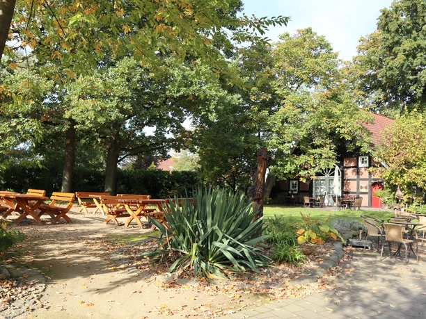 Biergarten Ein idyllischer Biergarten mit Holztischen und Stühlen im Schatten großer, grüner Bäume.