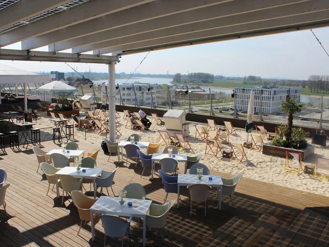 MonBerg mit Terrasse und Strand am Rhein Strandbar mit Sand, Sonnenliegen und Tischen auf einer Terrasse am Rhein unter einem offenen Dach.