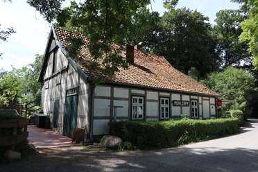 502B3A35-C8D4-4E78-8E1C-BA2390218E7E.jpeg Ein traditionelles Fachwerkhaus mit Ziegeldach, umgeben von Bäumen, steht am ruhigen Straßenrand.