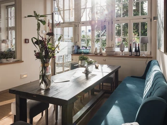 Genusswerkstatt interior 2 Heller Raum mit großem Holztisch, blauen Polsterbänken und Blumen, Sonnenlicht durchflutet Fenster.