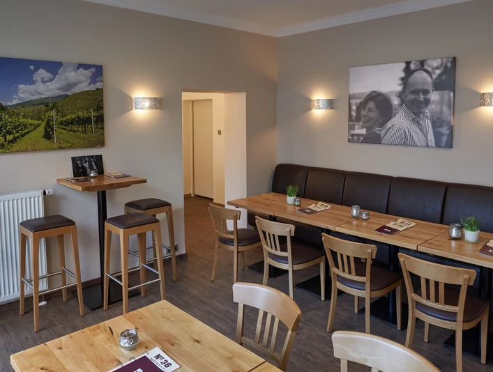 Gemütlicher Innenraum einer Weinbar mit Holztischen, Barhockern, Wandbildern von Weinbergen und Personen.