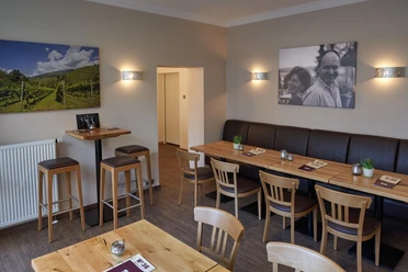 Weinbar 36_ Raum 1 Gemütlicher Innenraum einer Weinbar mit Holztischen, Barhockern, Wandbildern von Weinbergen und Personen.
