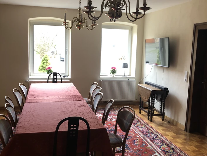 Langes Esszimmer mit Holzfußboden und großem Tisch, umgeben von Stühlen. Kronleuchter und Teppich betonen das Ambiente.