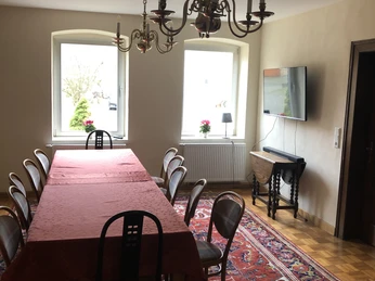 Esszimmer.JPG Langes Esszimmer mit Holzfußboden und großem Tisch, umgeben von Stühlen. Kronleuchter und Teppich betonen das Ambiente.