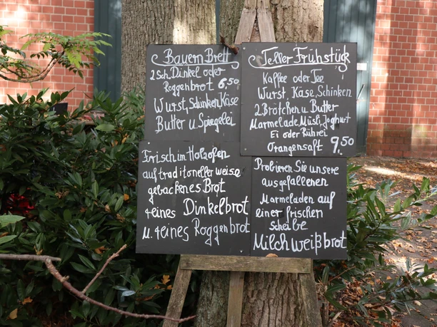Speisenangebot Tafel in Natur: Menüs mit Bauernfrühstück und frischem Brot; umgeben von grünen Sträuchern.