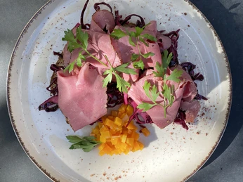 Anker_Villa Ein Teller mit rosa Roastbeef, Rotkraut und gelben Würfeln, dekoriert mit frischen Petersilienblättern.
