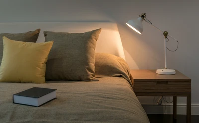 Musterbild Übernachtung Gemütliches Schlafzimmer mit beleuchtetem Nachttisch, Bett mit Kissen und Buch daneben.