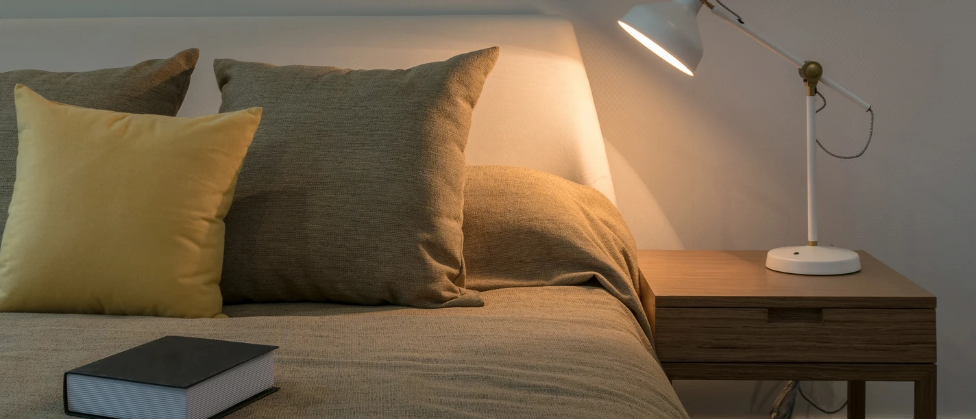 Musterbild Übernachtung Gemütliches Schlafzimmer mit beleuchtetem Nachttisch, Bett mit Kissen und Buch daneben.