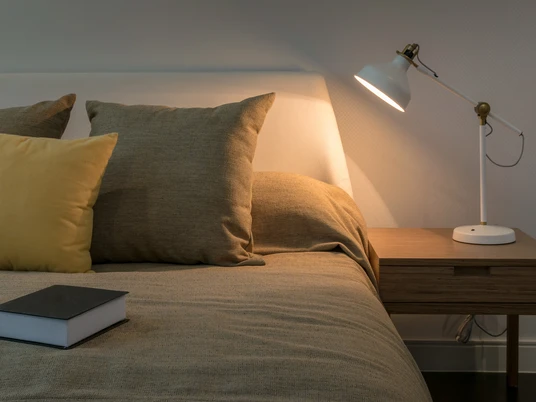 Musterbild Übernachtung Gemütliches Schlafzimmer mit beleuchtetem Nachttisch, Bett mit Kissen und Buch daneben.