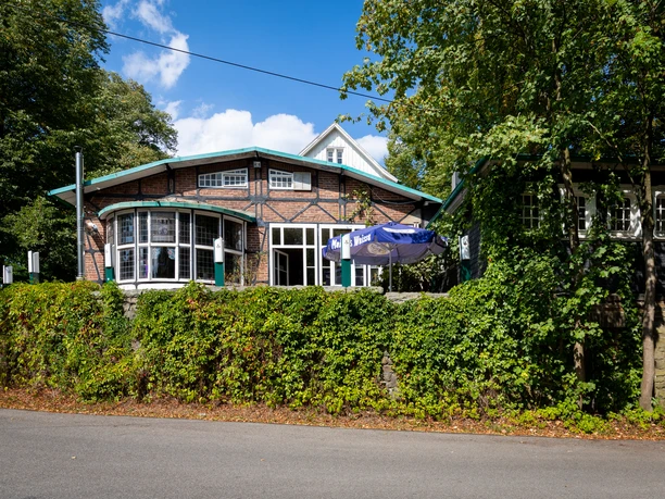 Haus Eifgen Rückansicht Historisches Gebäude mit Fachwerk, großer Fensterfront und Sonnenschirm, umgeben von dichter Vegetation.