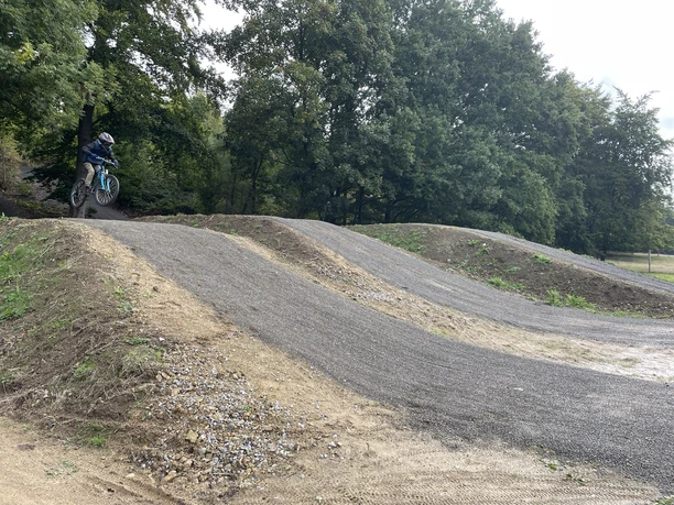 Ein Mountainbiker springt über einen Hügel im Bikepark mit dicht bewaldetem Hintergrund.