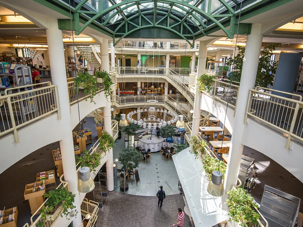 Stadtbibliothek Gütersloh Heller, moderner Innenraum eines mehrstöckigen Gebäudes mit Glasdach und begrünten Balkonen.
