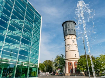 Glasfassade eines modernen Gebäudes und ein historischer Wasserturm bei strahlend blauem Himmel.