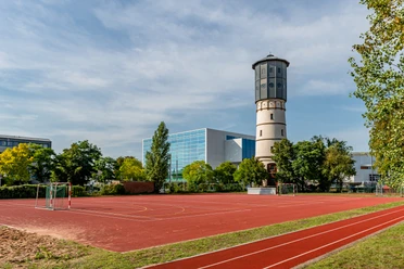 Historischer Wasserturm neben einem Sportplatz mit Basketballkörben und blauen Gebäude im Hintergrund.