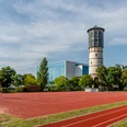 Historischer Wasserturm neben einem Sportplatz mit Basketballkörben und blauen Gebäude im Hintergrund.