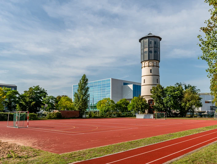 Wasserturm-Theater-GT-M.Wallenfang (1).jpg Historischer Wasserturm neben einem Sportplatz mit Basketballkörben und blauen Gebäude im Hintergrund.