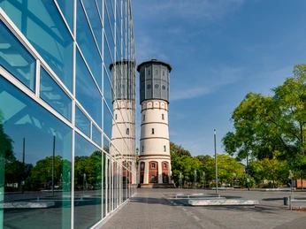 Wasserturm Gütersloh Moderner Glasbau spiegelt historischen Wasserturm wider, daneben grüne Bäume und blauer Himmel.