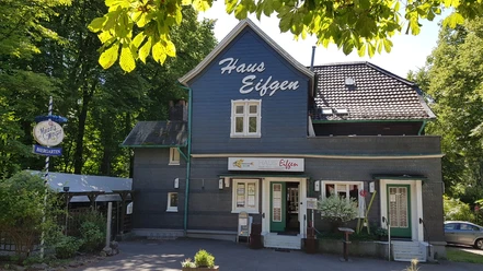 Haus Eifgen Gasthaus in einem blauen Fachwerkhaus, umgeben von grünen Bäumen, mit dem Schild "Haus Eifgen".