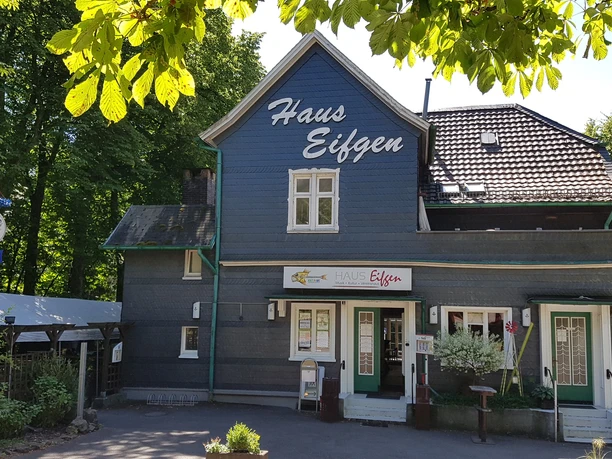 Haus Eifgen Gasthaus in einem blauen Fachwerkhaus, umgeben von grünen Bäumen, mit dem Schild "Haus Eifgen".