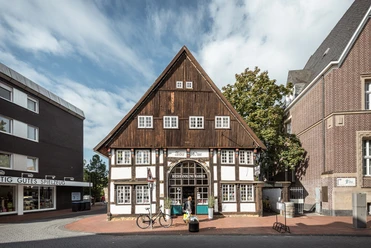 Ein historisches Fachwerkhaus mit dunklem Holzdach und weißer Fassade, umgeben von modernen Gebäuden.
