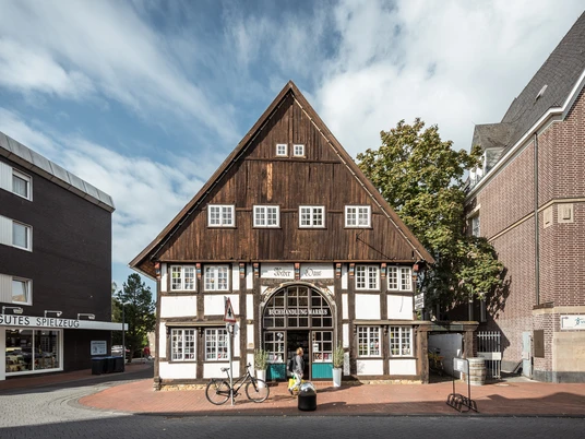 Ein historisches Fachwerkhaus mit dunklem Holzdach und weißer Fassade, umgeben von modernen Gebäuden.