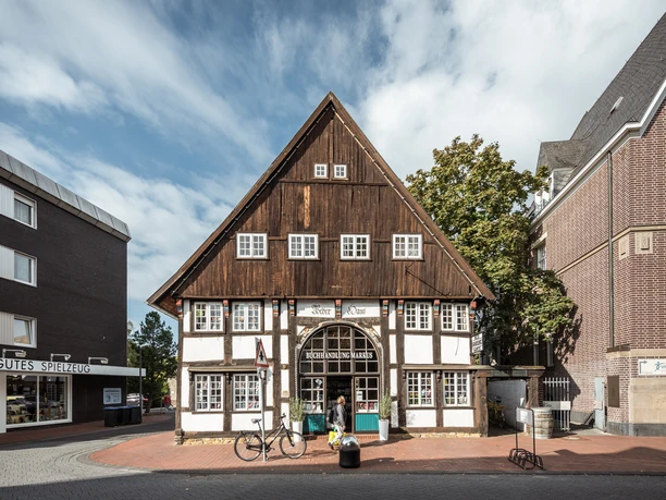 Weberhaus Ein historisches Fachwerkhaus mit dunklem Holzdach und weißer Fassade, umgeben von modernen Gebäuden.