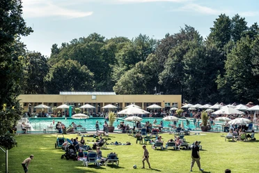 Parkbad Gütersloh Menschen entspannen und genießen die Sonne auf einer grünen Liegewiese vor einem großen Freibad.