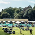 Parkbad Gütersloh Menschen entspannen und genießen die Sonne auf einer grünen Liegewiese vor einem großen Freibad.