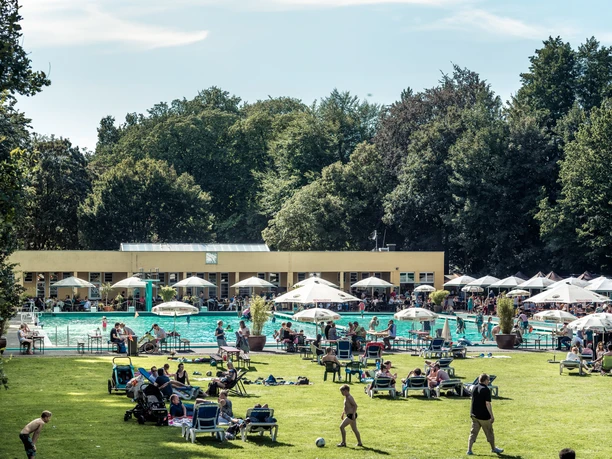 Parkbad Gütersloh Menschen entspannen und genießen die Sonne auf einer grünen Liegewiese vor einem großen Freibad.