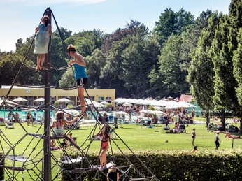 Klettergerüst Parkbad Gütersloh Kinder klettern auf einem großen Klettergerüst im Freien, umgeben von grünen Bäumen und einem Poolbereich.