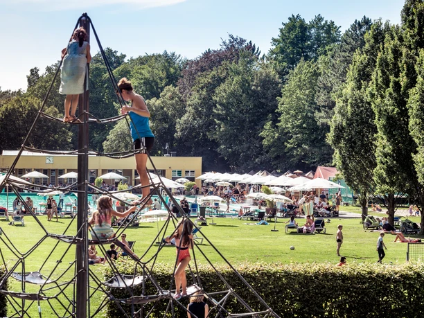 Klettergerüst Parkbad Gütersloh Kinder klettern auf einem großen Klettergerüst im Freien, umgeben von grünen Bäumen und einem Poolbereich.