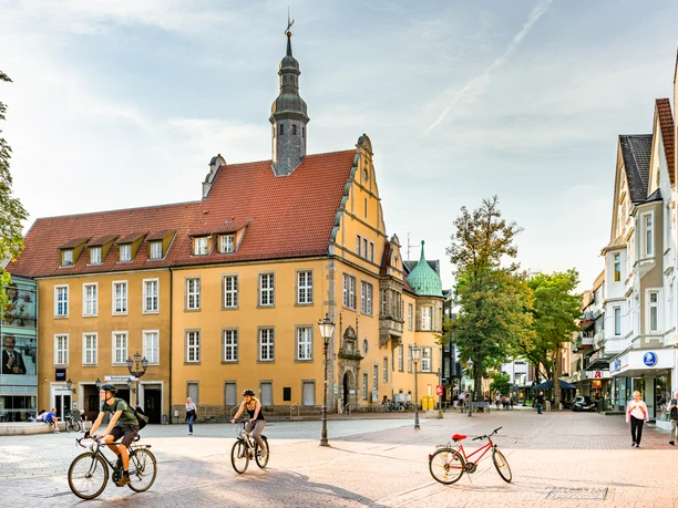 Historisches Gebäude mit spitzem Turm, umgeben von Radfahrern und Fußgängern auf gepflastertem Platz.