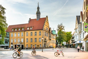 Altes Amtsgericht Gütersloh Historisches Gebäude mit spitzem Turm, umgeben von Radfahrern und Fußgängern auf gepflastertem Platz.