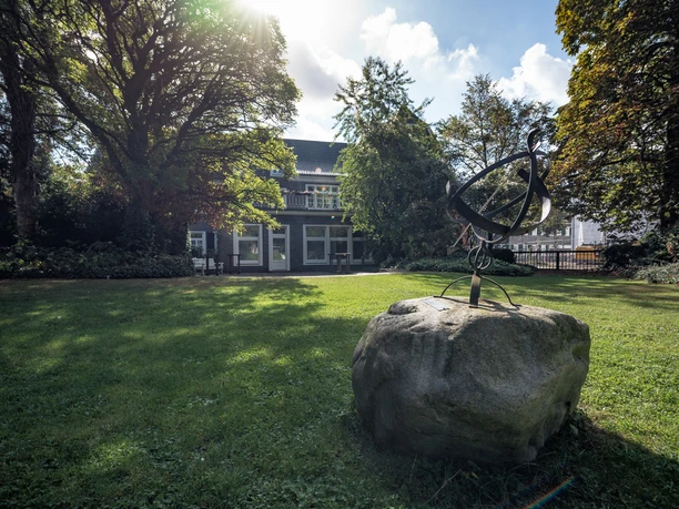 Ein Herrenhaus mit grauer Fassade, umgeben von Bäumen und Rasen, im Vordergrund ein abstrakte Skulptur.