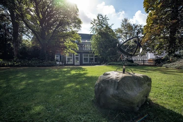 Villa Bartels Ein Herrenhaus mit grauer Fassade, umgeben von Bäumen und Rasen, im Vordergrund ein abstrakte Skulptur.