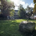 Villa Bartels Ein Herrenhaus mit grauer Fassade, umgeben von Bäumen und Rasen, im Vordergrund ein abstrakte Skulptur.