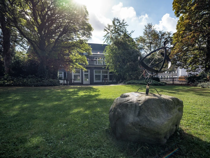 Villa Bartels Ein Herrenhaus mit grauer Fassade, umgeben von Bäumen und Rasen, im Vordergrund ein abstrakte Skulptur.