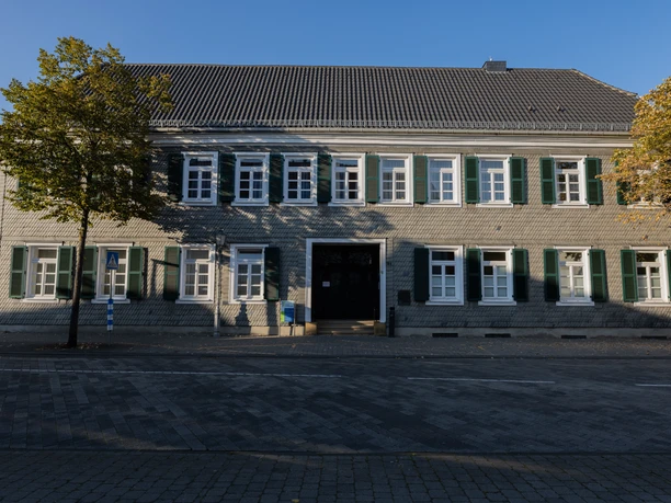 Zweistöckiges historisches Gebäude in Backsteinoptik mit grünen Fensterläden und flankierenden Bäumen.