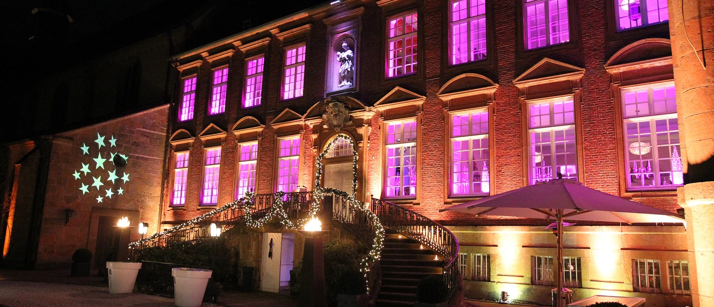 Alte Abtei im Lichterglanz von der Klosterpforte.jpg Historisches Gebäude bei Nacht mit violett beleuchteten Fenstern und festlich geschmücktem Eingang.
