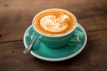 Eine hellgrüne Tasse mit Cappuccino, verziert mit Latte Art, steht auf einem hölzernen Tisch.
